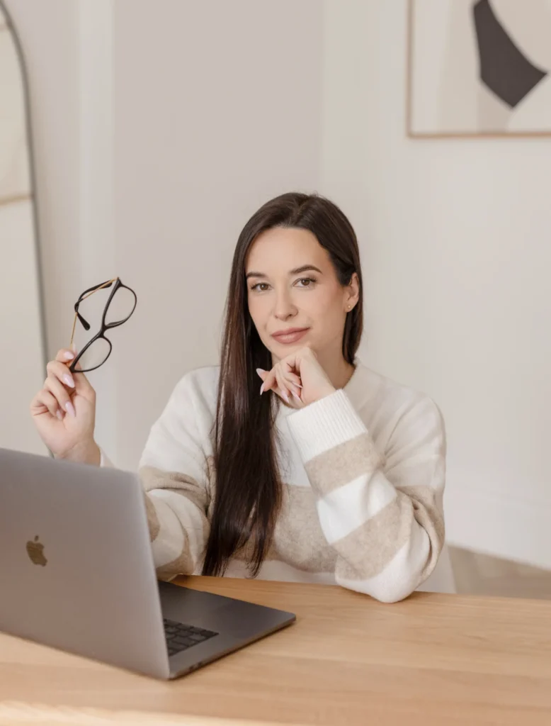 Młoda kobieta o długich ciemnych włosach siedzi uśmiechnięta przy biurku, na którym stoi szary laptop.